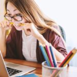 ノートパソコンの画面を見ながら鉛筆を噛む眼鏡の女性。緊張やもどかしさのイメージ